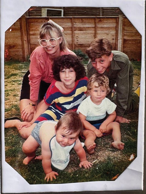 A family portrait featuring five smiling individuals of various ages, from baby to adult, posing outdoors.