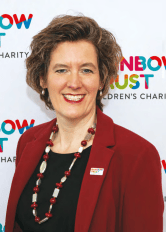 A smiling woman in a red jacket with a Rainbow Trust Children's Charity logo in the background.