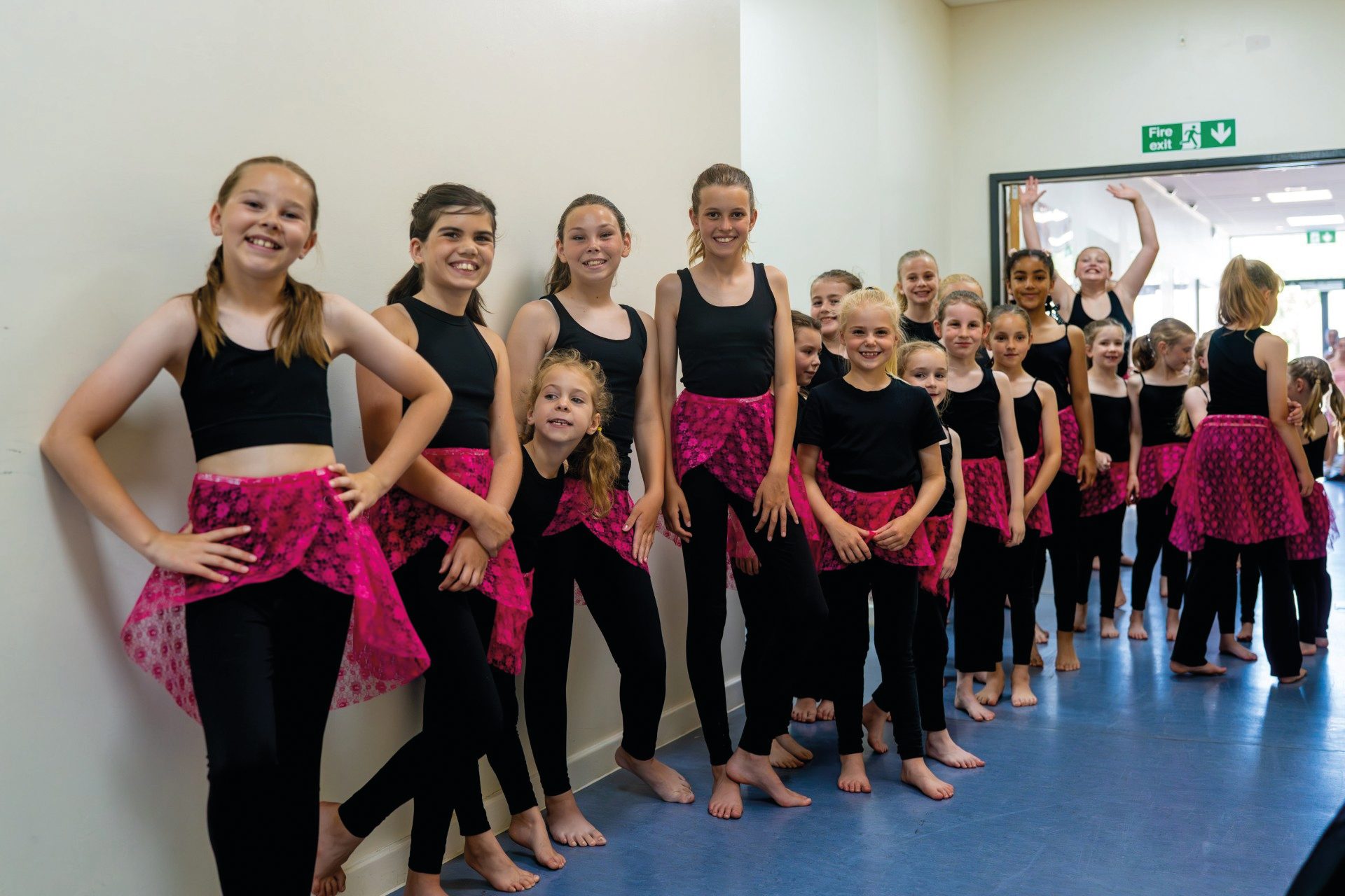 A group of smiling girls in black and pink dance costumes, standing barefoot indoors.