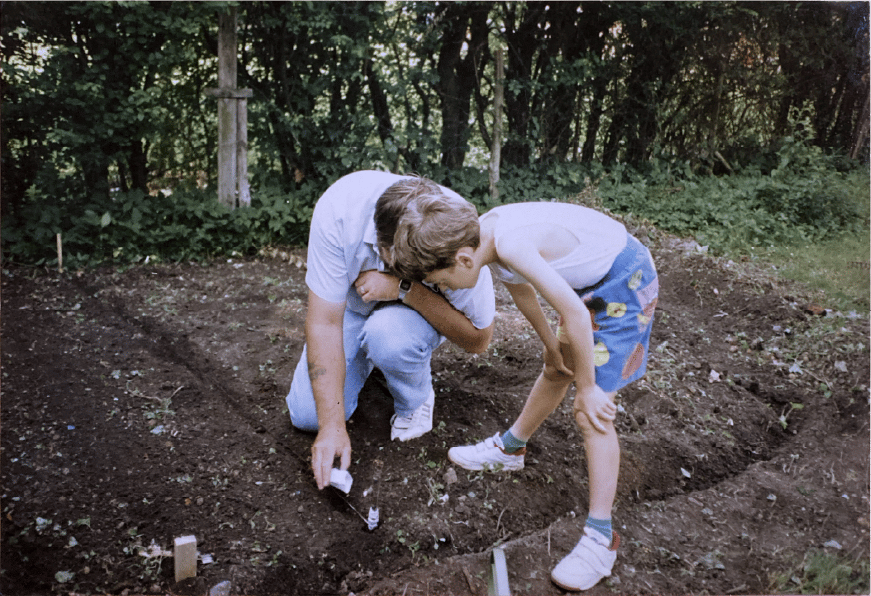 A man and boy planting seeds in a garden bed, with green foliage in the background.
