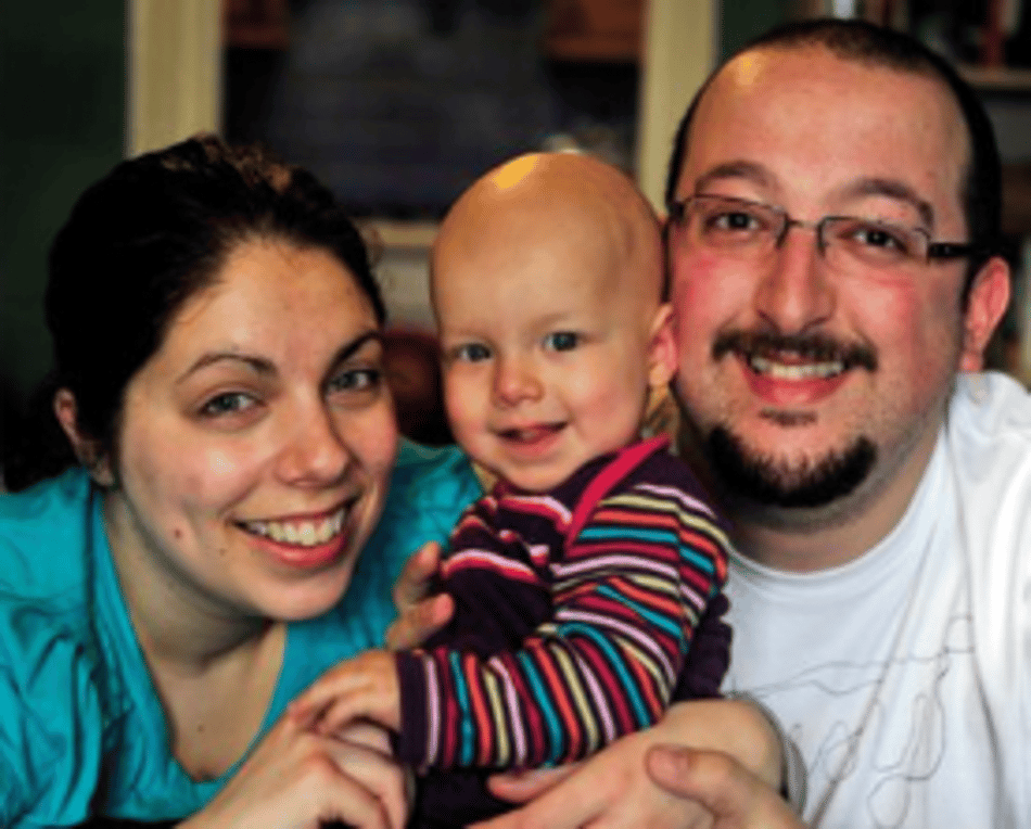 A smiling family portrait featuring a mother, father, and their bald baby, looking happy together.