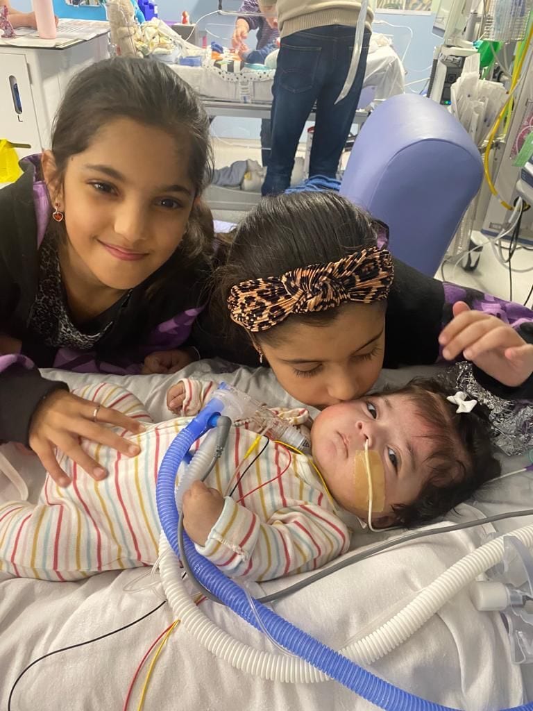 Two girls lovingly interact with a baby connected to medical equipment in a hospital bed.
