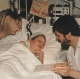 Two adults attentively comfort a child in a hospital bed, reading from a document together.