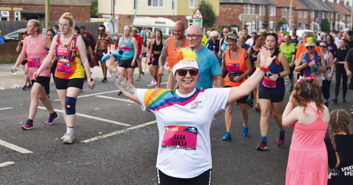 Smiling woman in rainbow shirt with arms out, leading a crowd at an outdoor event.