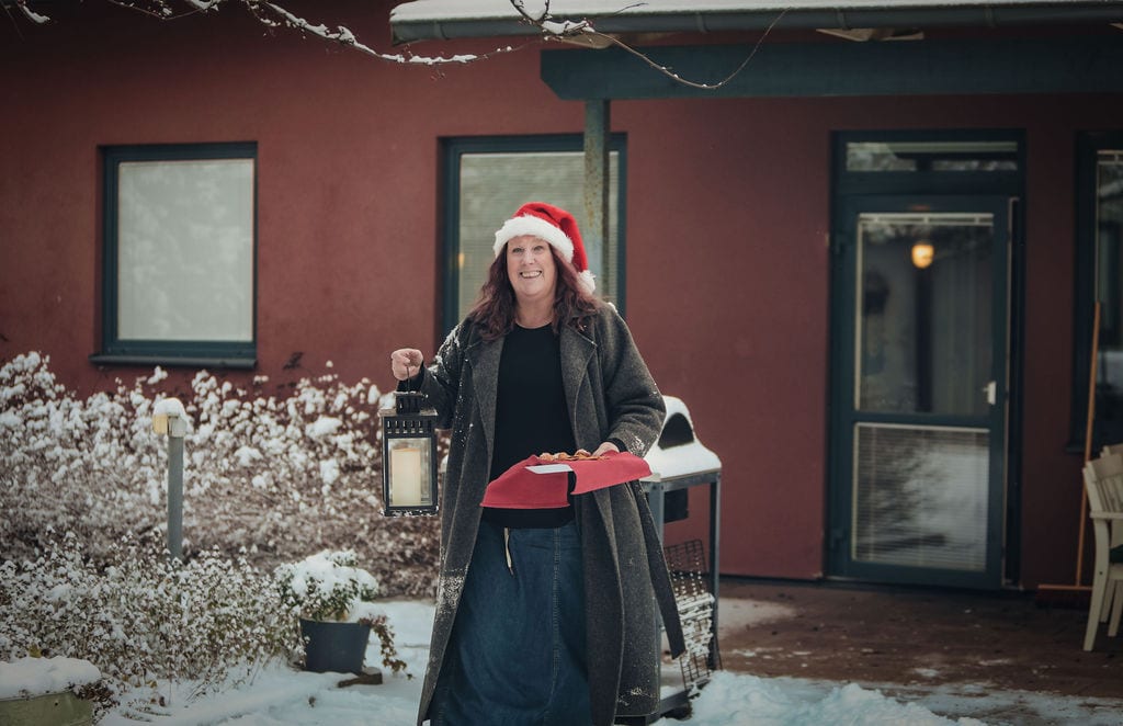 Woman in Santa hat holding a lantern and cookies in the snow outside a red house.