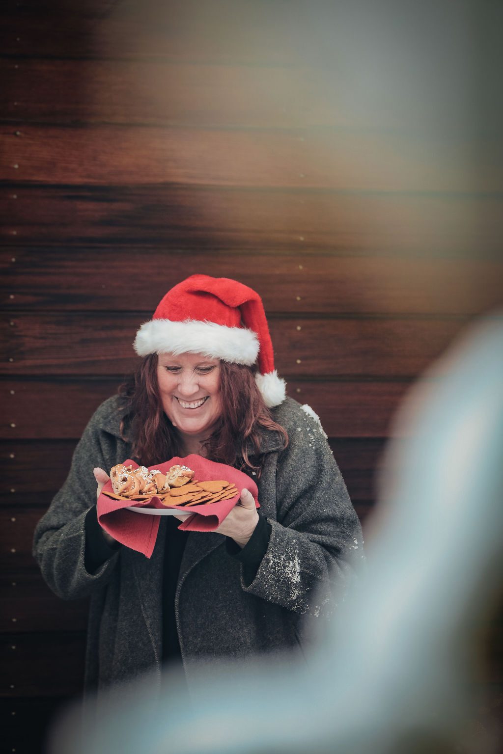 Smiling woman in Santa hat holds a plate of Christmas baked goods, snow on her coat.