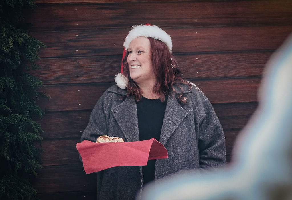 Happy woman in a Santa hat smiles, holding pastries on a red cloth against a wooden wall and pine tree.