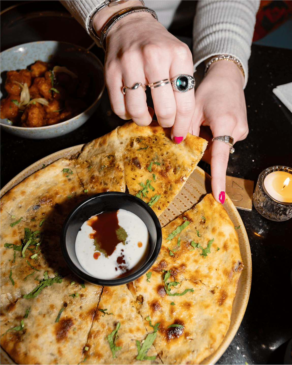 Close-up of hands taking naan from a platter with dip, curry, and candlelight.