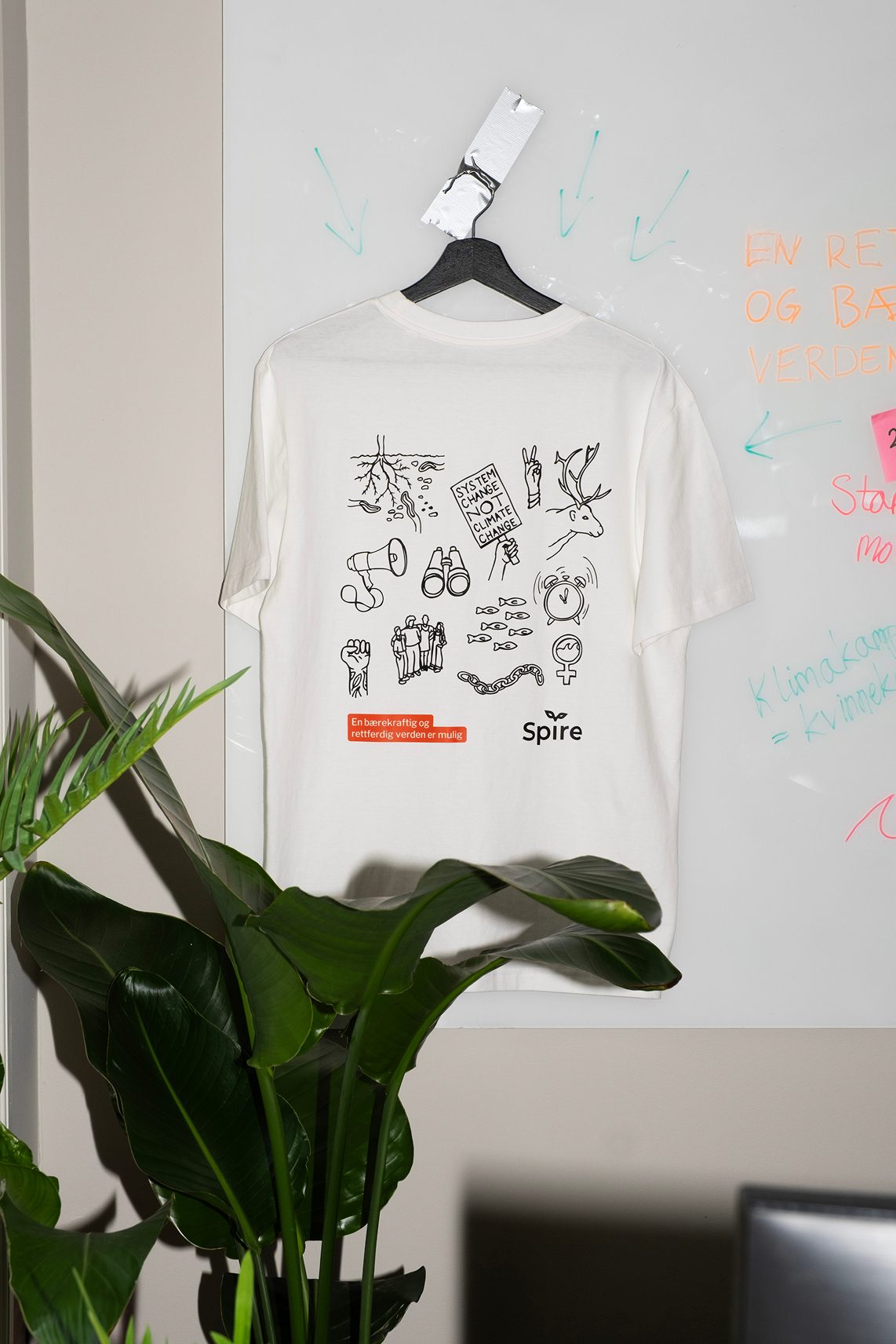 White t-shirt with activist illustrations and "System Change" text hangs on a whiteboard, plant in front.