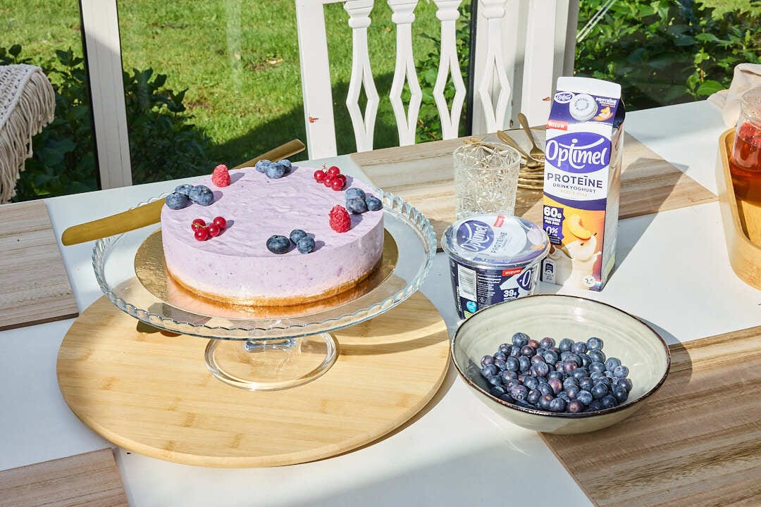Blueberry cheesecake with berries, bowl of blueberries, and Optimel protein products outdoors.