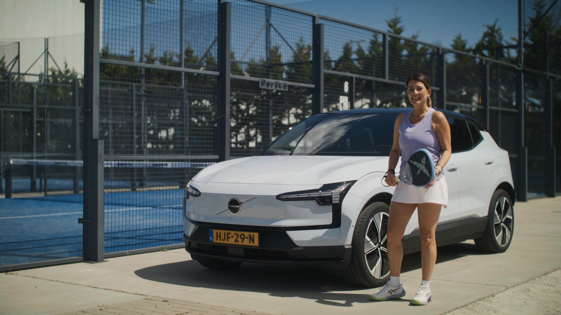 A woman holds a padel racket next to a white Volvo car near a padel court.
