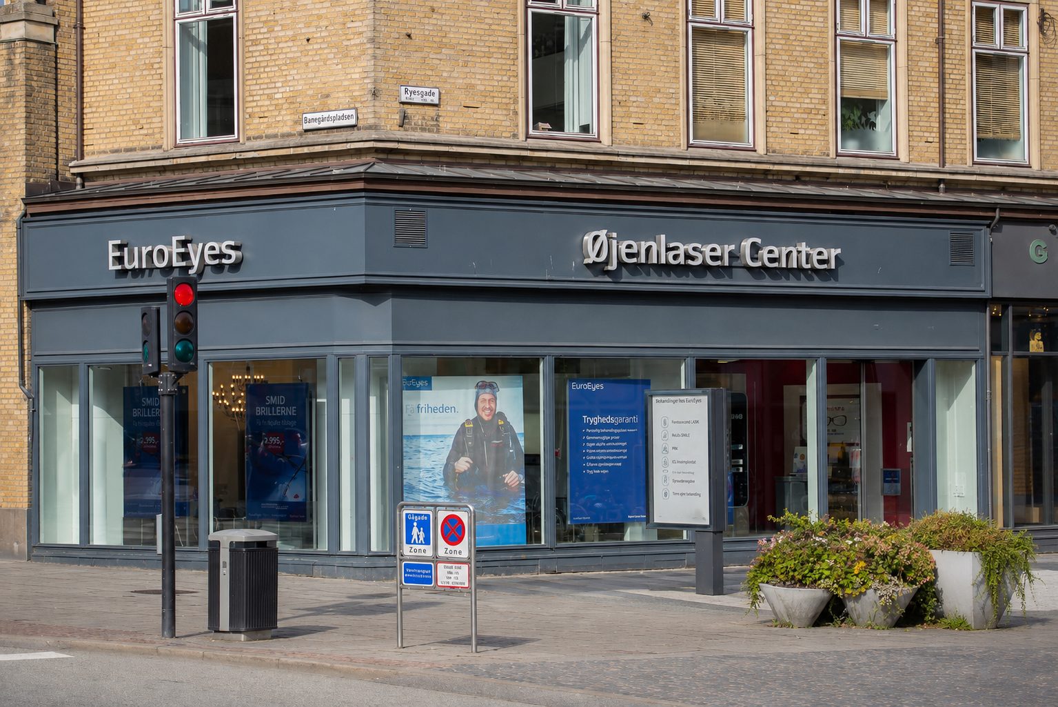 Daytime view of EuroEyes and Øjenlaser Center shops at a street corner with a traffic light and posters.