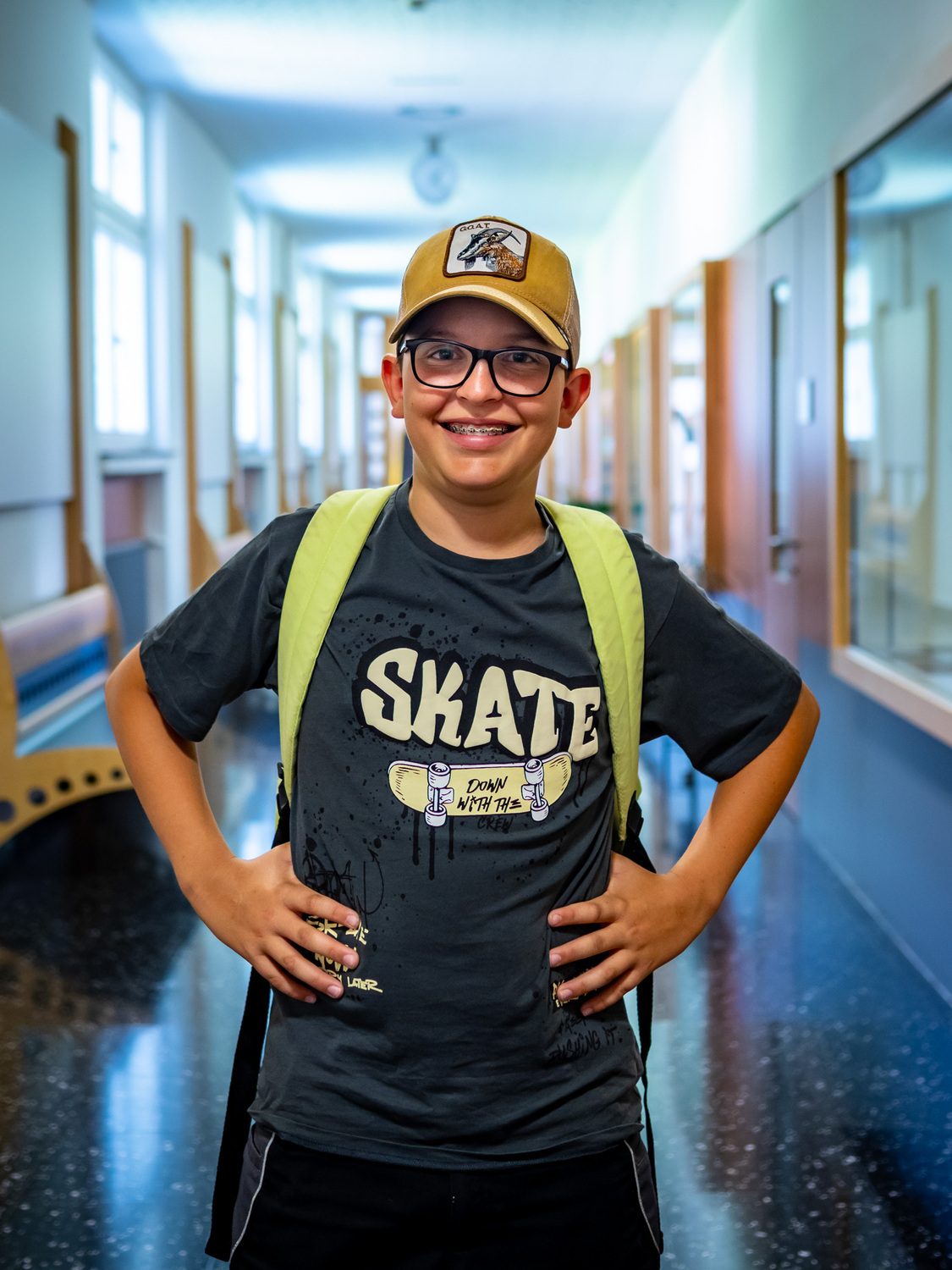 Smiling boy with glasses, braces, a "SKATE" t-shirt, and backpack stands in a school hallway.