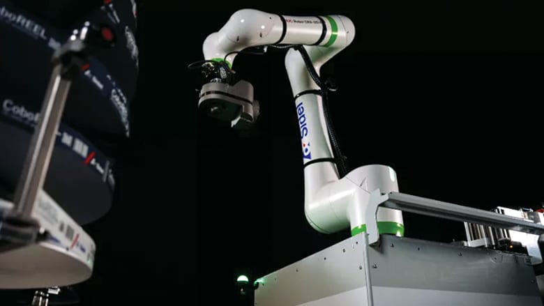 White and green robotic arm working with "CoboREEL" industrial equipment in a dark setting.