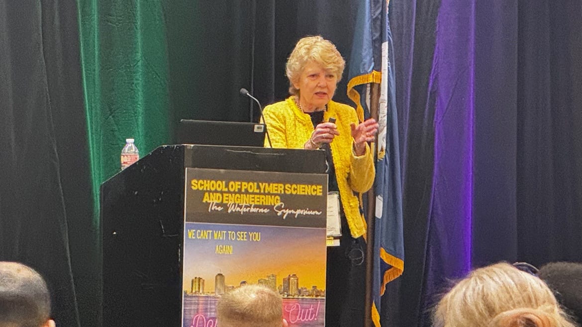 An older woman in a yellow jacket speaks at a podium with "School of Polymer Science" signage.