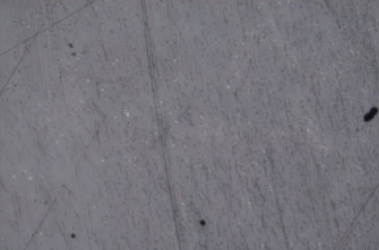 Grey surface with fine scratches and scattered dark specks.
