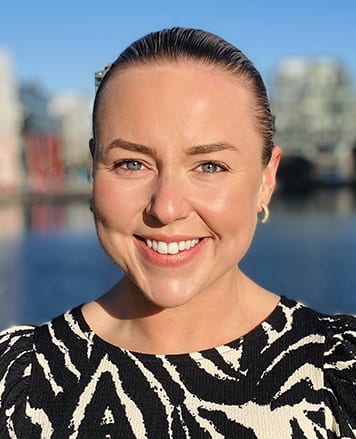 A smiling woman with her hair pulled back, wearing a black and white patterned top, outdoors.