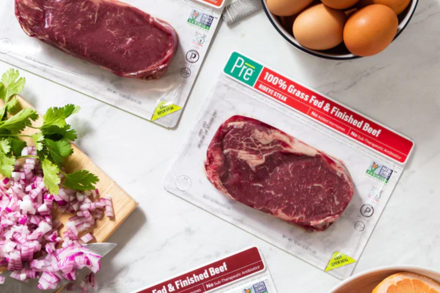 Packaged grass-fed ribeye steaks with chopped red onions, cilantro, and eggs on a marble counter.