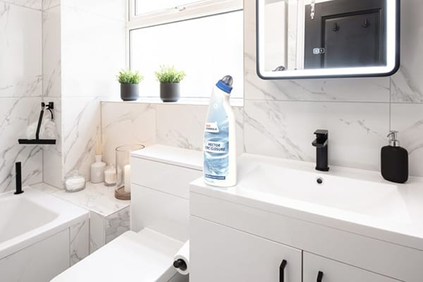 A clean, modern bathroom featuring white marble-effect tiles, a vanity, toilet, and bathtub with a cleaning product bottle.