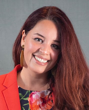 A smiling woman with long red hair, wearing a floral top and an orange blazer.