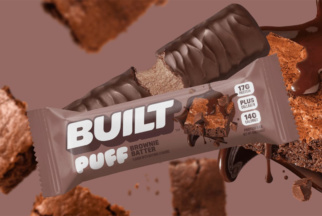 Built Puff Brownie Batter protein bar, both packaged and bitten, surrounded by brownie pieces and chocolate drips.
