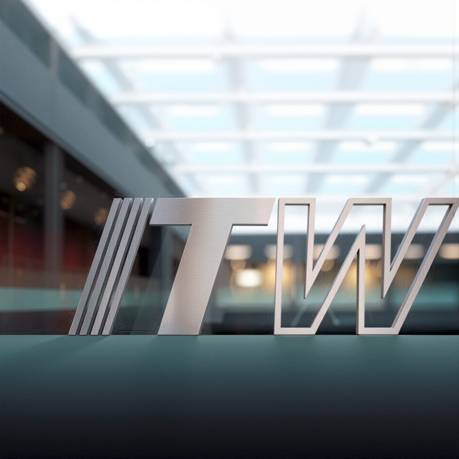 A close-up shot of the metallic, brushed aluminum 'TIW' logo with three diagonal lines on the left, against a blurred modern architectural background with a glass ceiling.