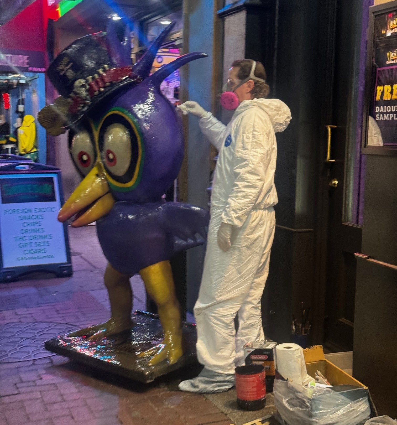 Person in a hazmat suit and respirator painting a large purple bird statue with a top hat.