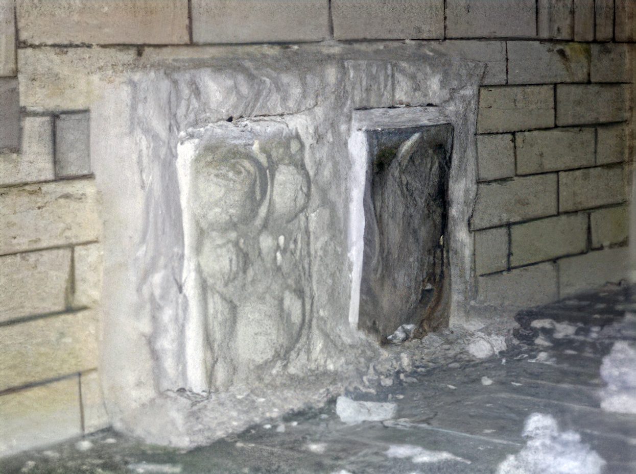 Worn stone carving of figures next to a dark alcove in an ancient stone wall.