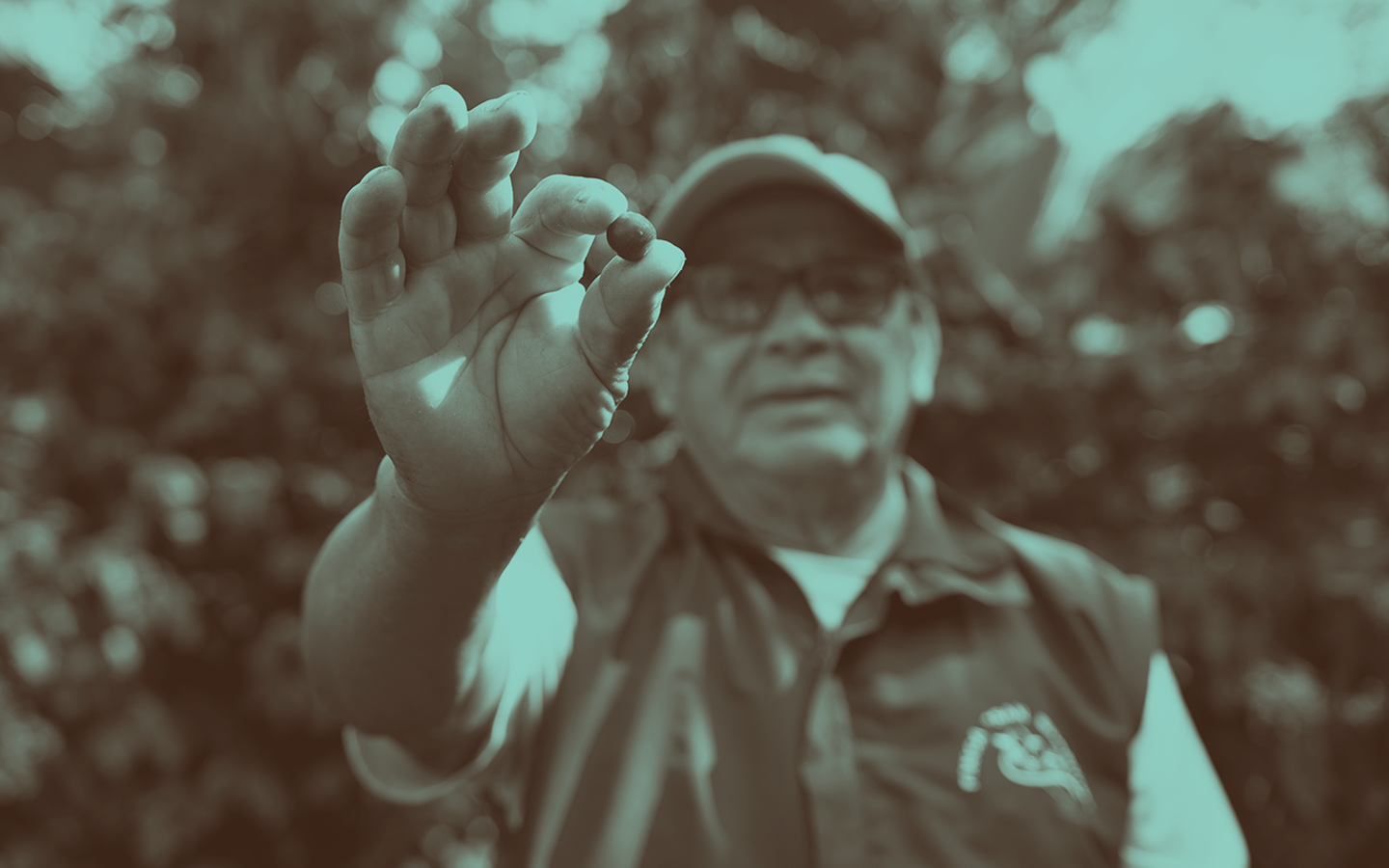 A hand in focus holds a dark berry or seed, with a blurred person in a cap in the background.