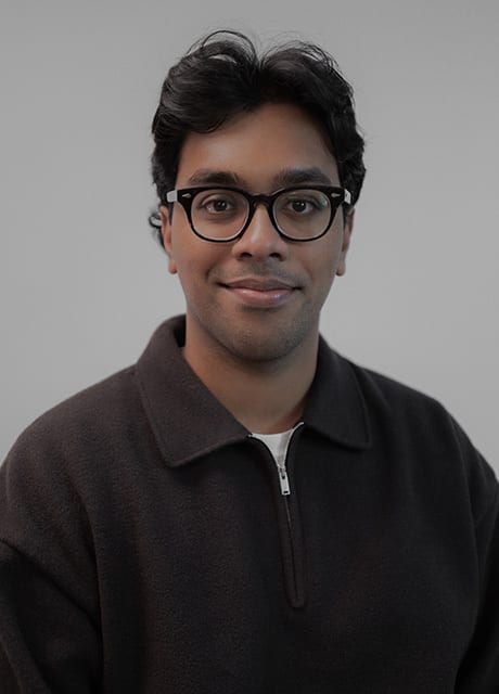 Smiling man with dark hair and glasses, wearing a dark brown top.