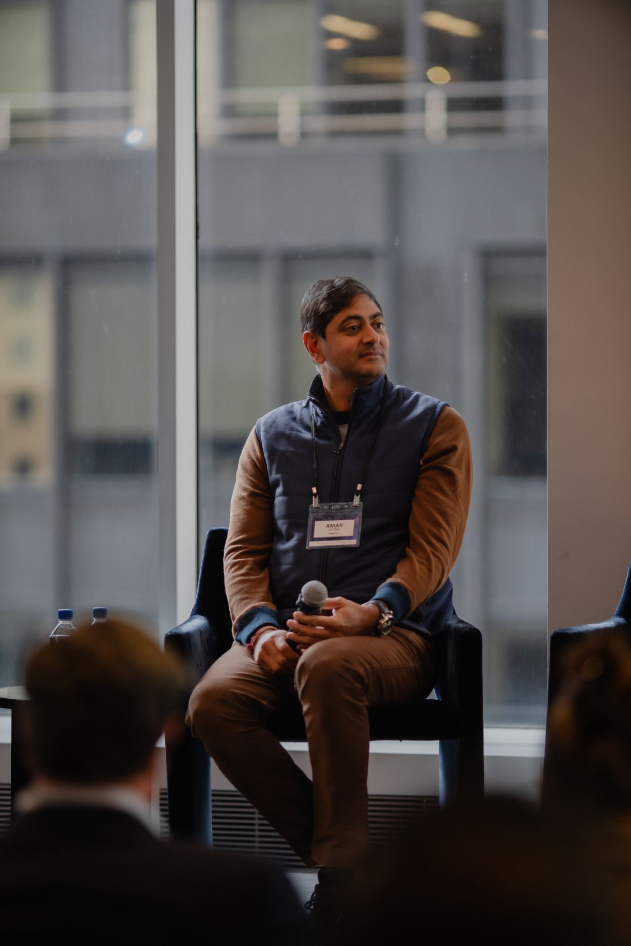 Amar, seated and holding a microphone, looks right with a city view through a window behind him.