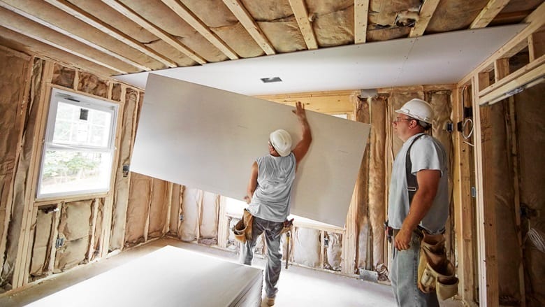 Two construction workers installing large drywall sheets on a ceiling in a room under renovation.