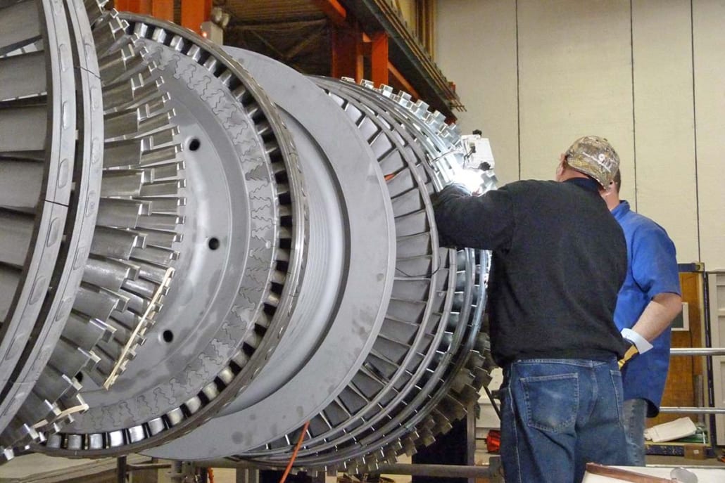 Two technicians work on assembling a large industrial turbine rotor with multiple stages of blades.