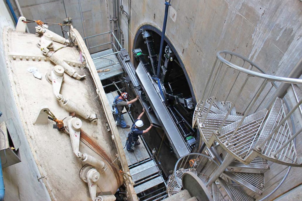 Two workers install a component into a large circular opening, flanked by heavy machinery and a spiral staircase.