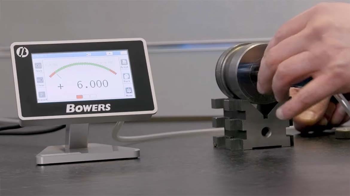 Hands measure a cylindrical part on a metal stand using a digital Bowers display showing 6.000.