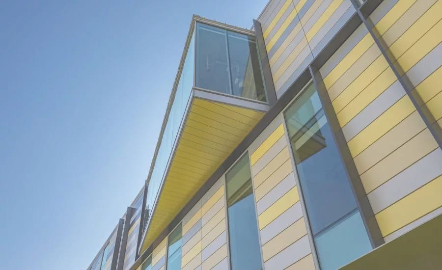 Modern building with yellow and grey striped facade, large windows, and a prominent glass skywalk under a clear sky.