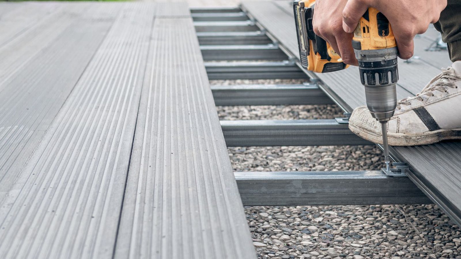 A person installing grey composite deck boards onto a metal frame using a power drill.