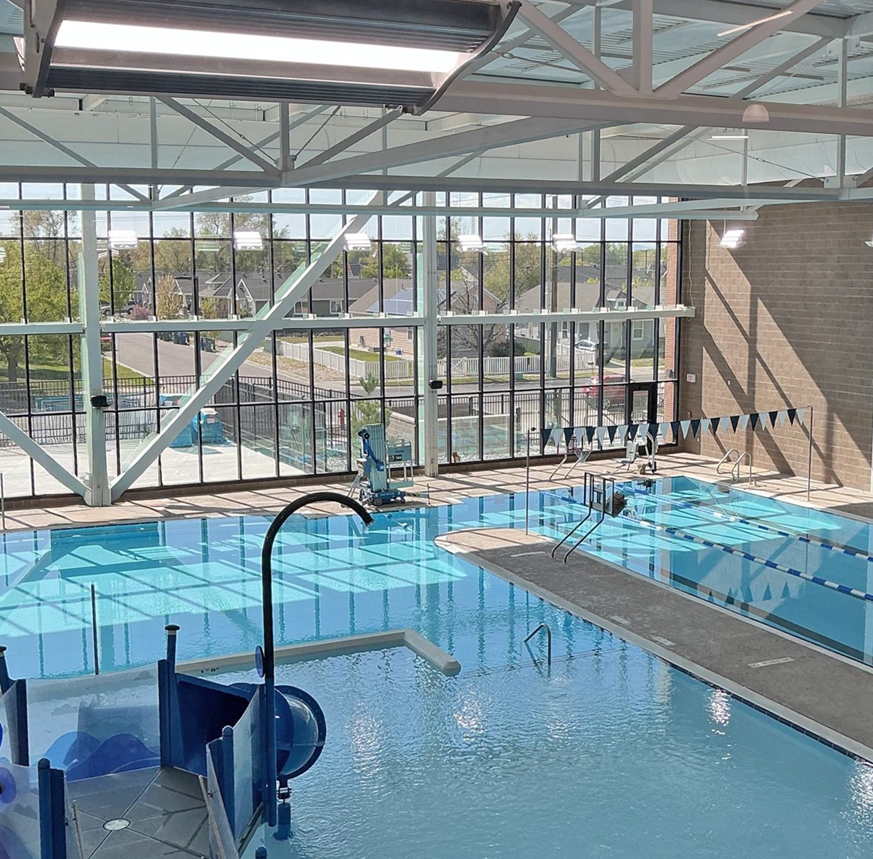Modern indoor swimming pool with large windows, lap lanes, and a children's slide play area.