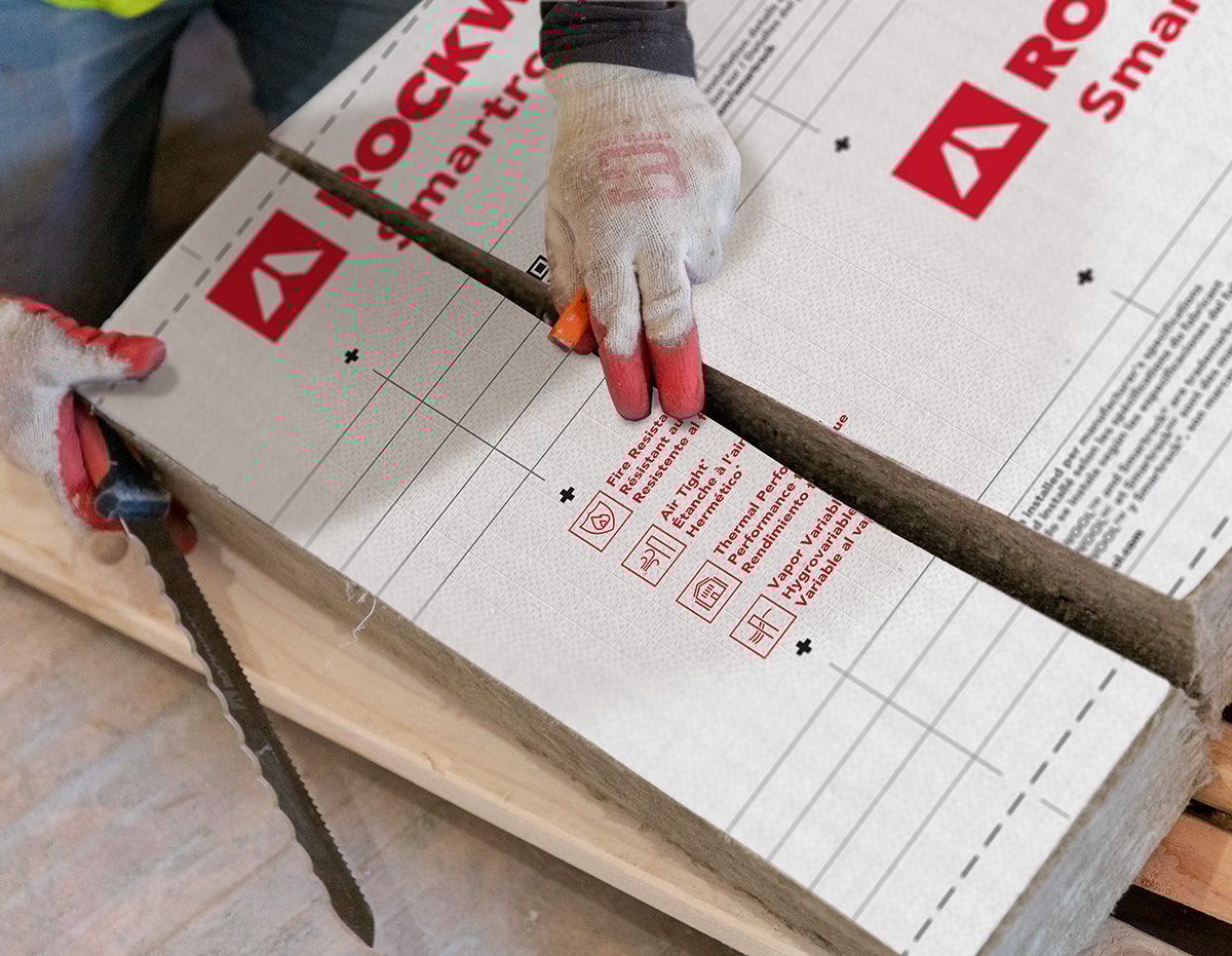 A worker's gloved hands are shown cutting Rockwool Smartro insulation with a saw blade.