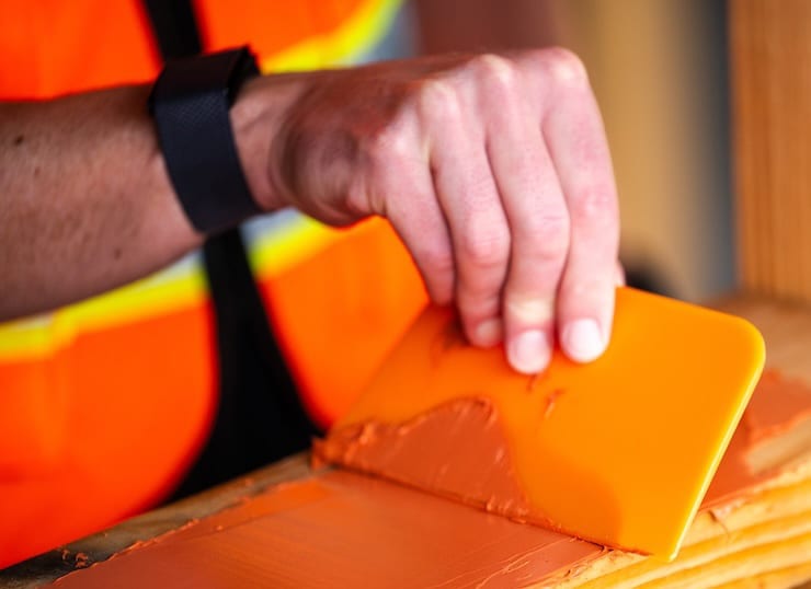 Hand in a safety vest using an orange squeegee to spread a reddish-brown paste.