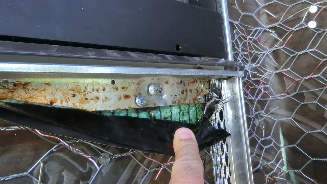Finger pointing at a corroded metal frame, green insulation, and wiring within a damaged appliance.