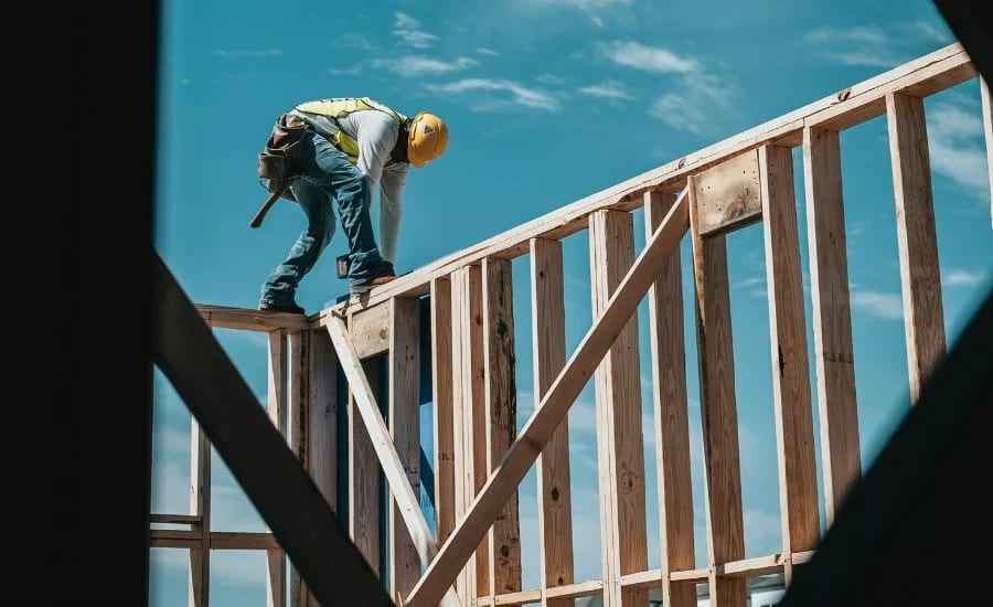 A construction worker wearing a hard hat and safety vest, working on the wooden frame of a house against a blue sky.