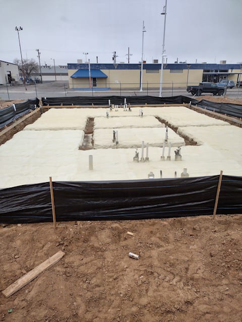 Construction foundation with foam insulation, plumbing pipes, and a black silt fence at a site.