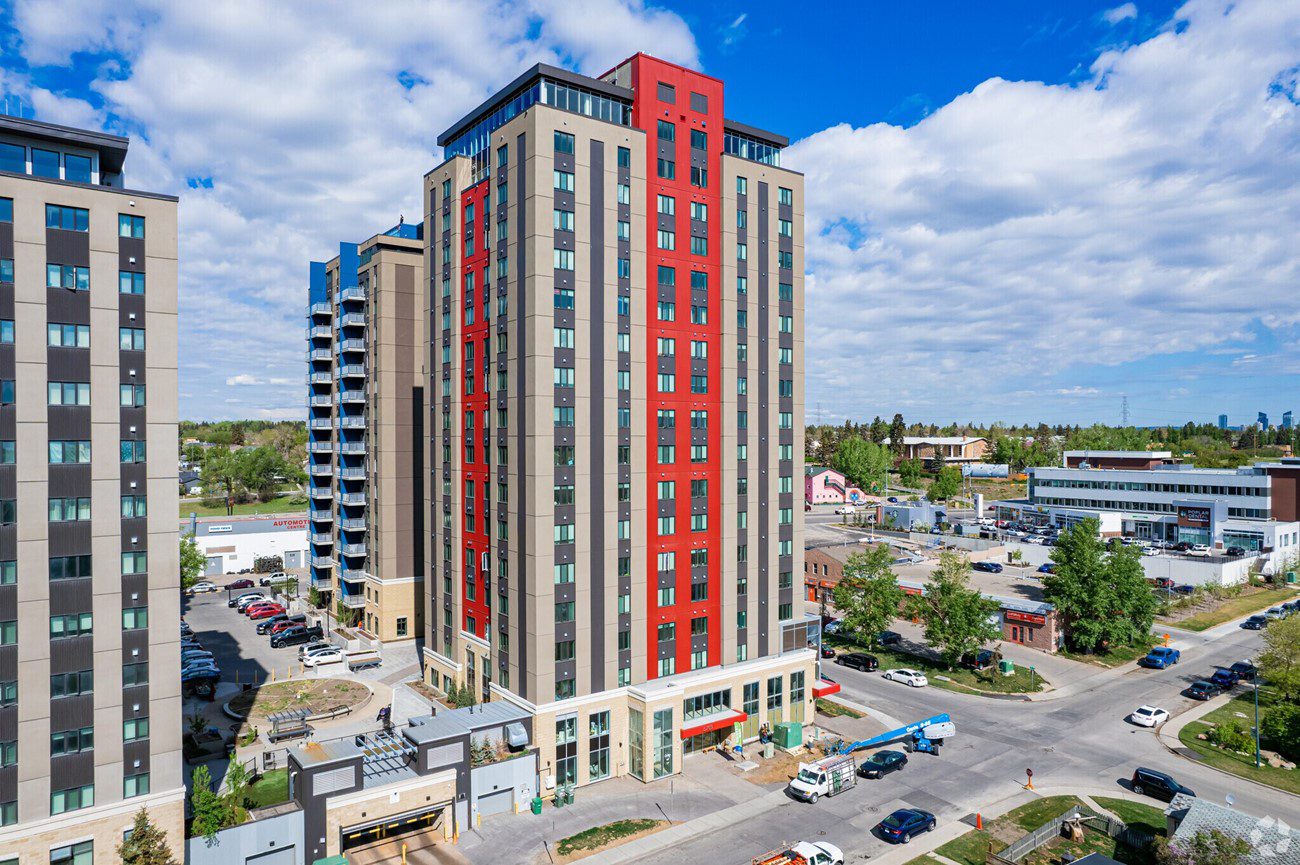 Modern high-rise buildings with red and grey panels, an urban streetscape, under a cloudy blue sky.