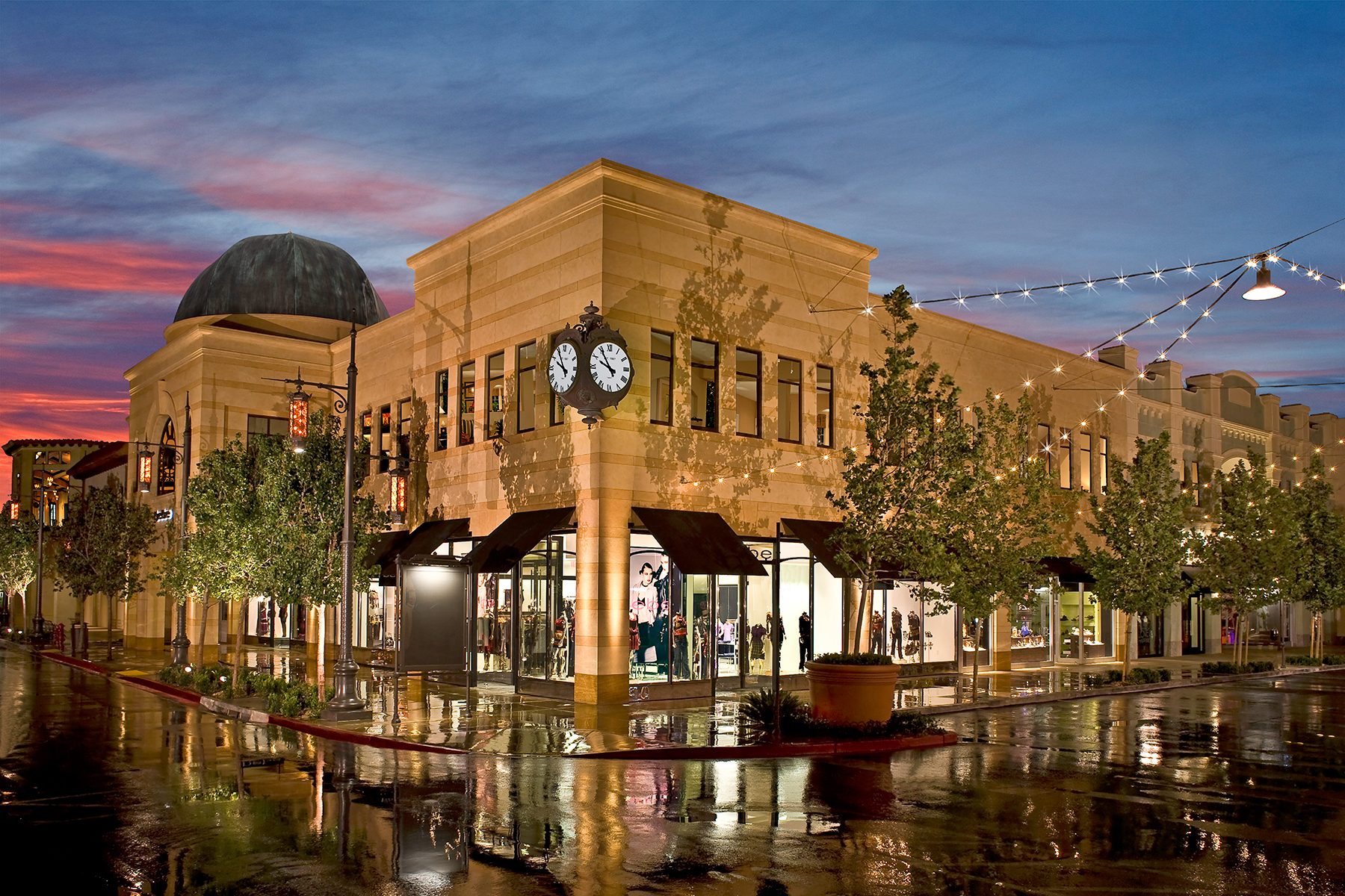 Illuminated shopping center at dusk with a corner clock, wet streets reflecting lights, and a colorful sky.