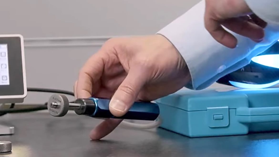 A person's hand holds a blue-handled tool with a metal disk, next to a display unit.