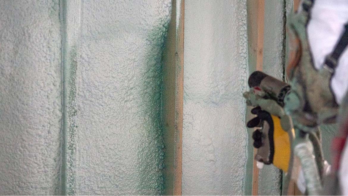 A worker's gloved hand applies spray foam insulation to wall studs, showing the foamy texture.