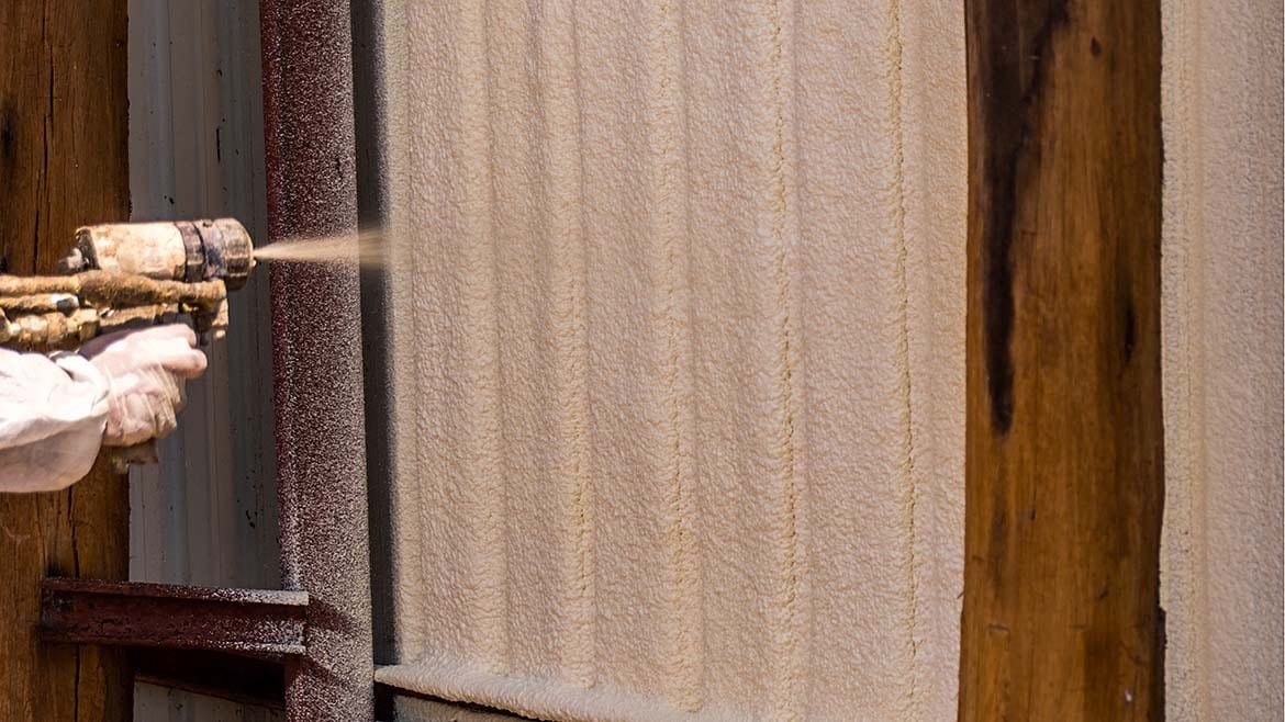 Hand spraying foam insulation onto a textured beige wall next to wooden beams.