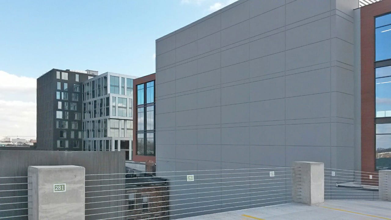 Modern grey and glass buildings, seen from a parking garage with numbered concrete posts and railings.