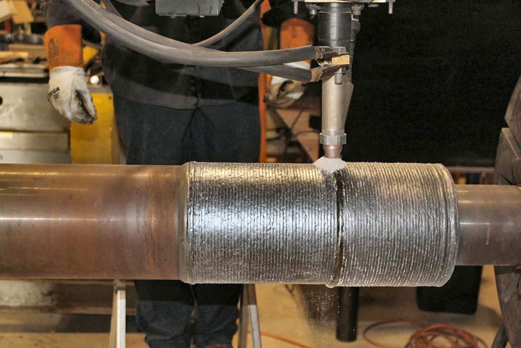A machine applies new material to a spinning metal shaft, creating a textured, built-up surface through welding.